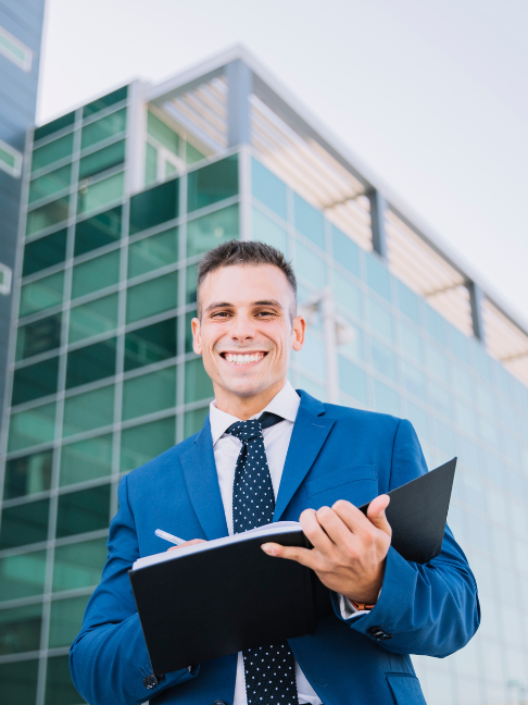 Representação do seguro empresarial. Homem sorrindo, de frente, anotando em um caderno de capa preta. Ao fundo, um prédio com janelas esverdeadas.