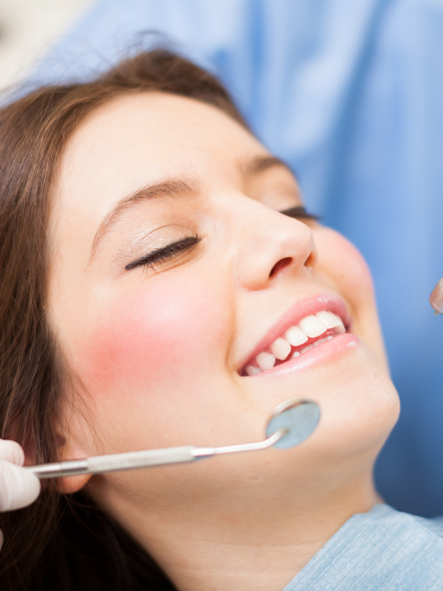 Imagem ilustrativa para o seguro odontológico PJ. Mulher sorrindo, com dentista ao fundo usando jaleco azul, enquanto segura um espelho bocal próximo à boca da paciente.